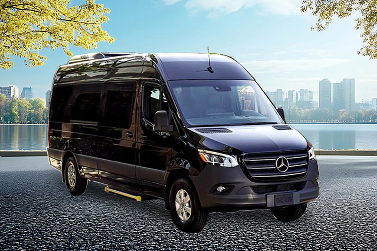 A black Mercedes-Benz Sprinter shuttle van parked beside a waterfront with trees and a city skyline behind it.