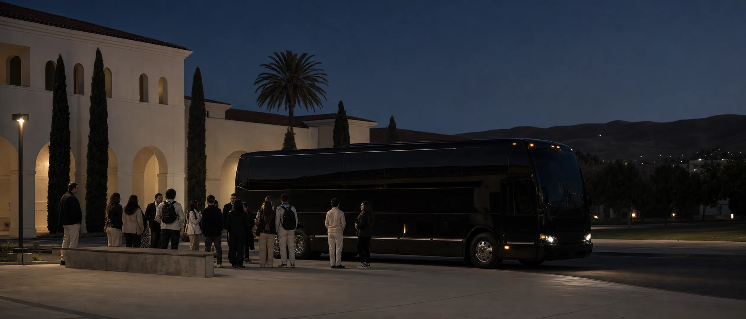 Students and chaperones gather beside a black motorcoach near Bakersfield College at dusk.